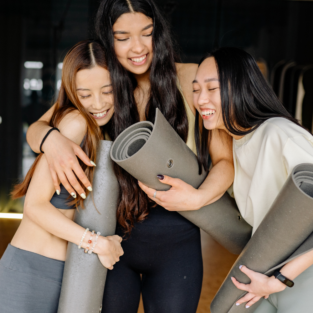 Photo of 3 women laughing and hugging, holding their rolled up yoga mats