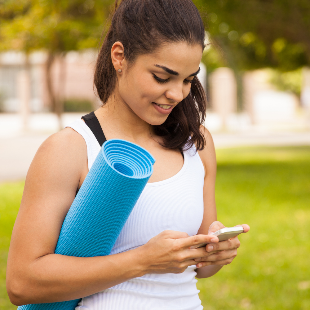 photo of woman holding a yoga mat and looking at her smart phone